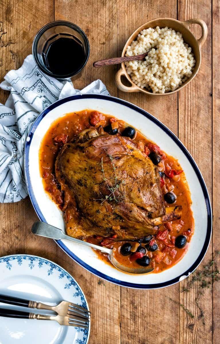 Tender slow cooker lamb shoulder, served in tomato sauce with black olives on an oval platter, accompanied by couscous, a glass of red wine, and a stack of plates with cutlery on a wooden table.
