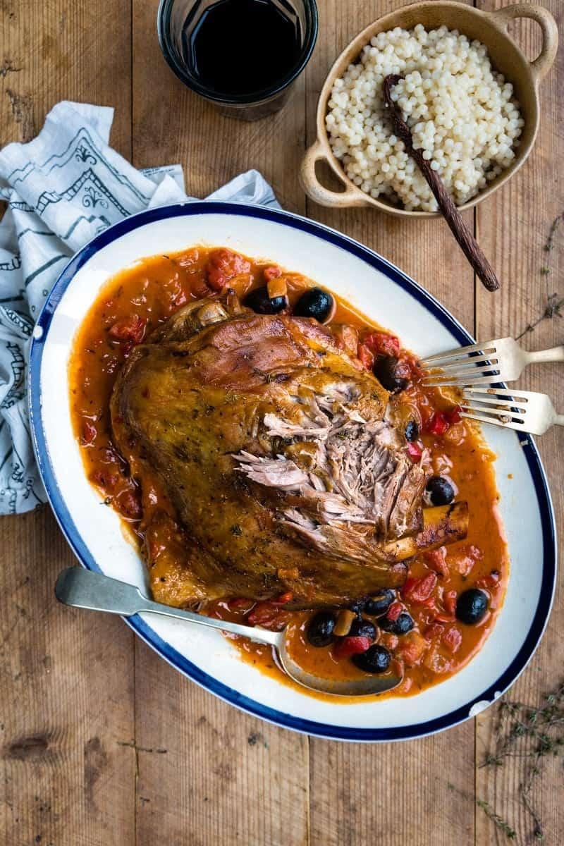 A One-pot Slow Cooked Lamb Shoulder sits on a large white platter in tomato sauce with black olives, with serving utensils beside it. Nearby, a bowl of couscous and a glass of red wine are on a wooden table.