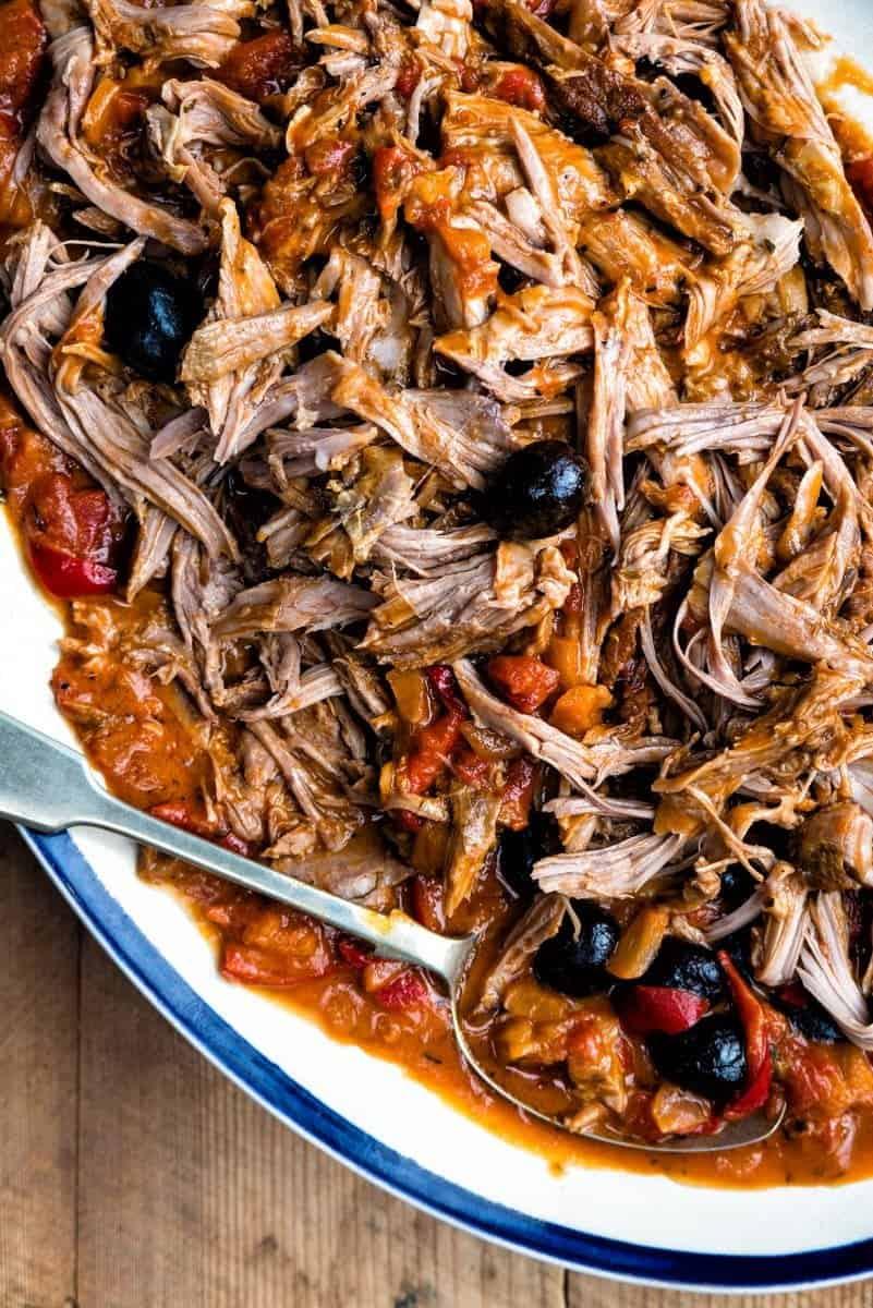 A close-up of shredded pork in a rich tomato-based sauce with black olives and red peppers, served on a white plate with a silver serving spoon—reminiscent of comforting one-pot slow cooked lamb shoulder meals.
