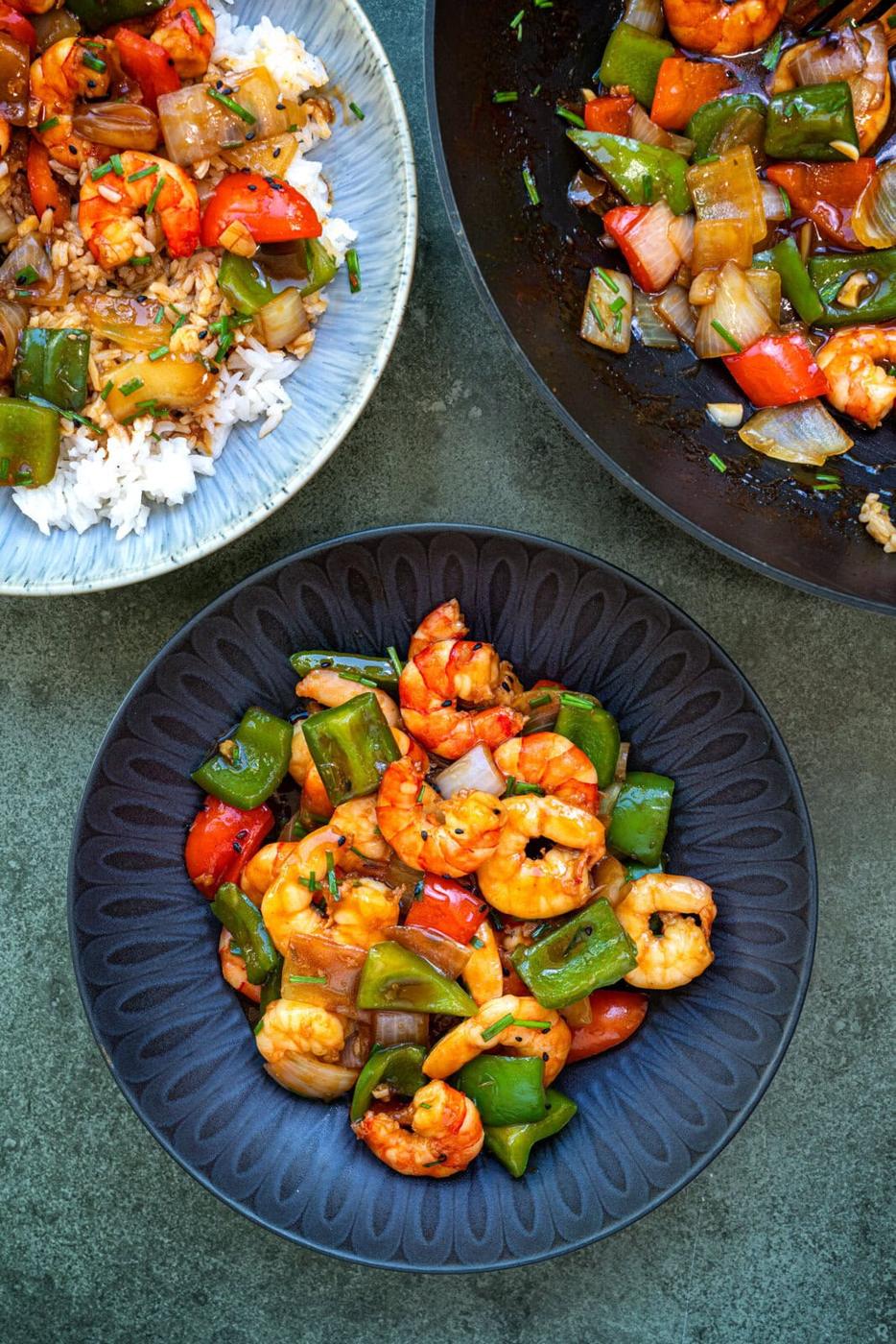 A dark plate filled with stir-fried shrimp, green bell peppers, red bell peppers, onions, and sauce. Nearby are a bowl with the stir-fry over rice and a wok with more of the colorful shrimp and vegetable dish.