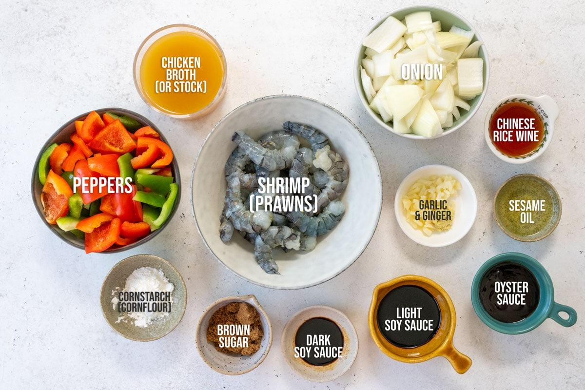 A top-down view of bowls containing ingredients for a stir-fry: raw shrimp in the center, surrounded by chopped onion, chopped peppers, chicken broth, garlic & ginger, oyster sauce, dark soy sauce, light soy sauce, sesame oil, rice wine, and brown sugar.