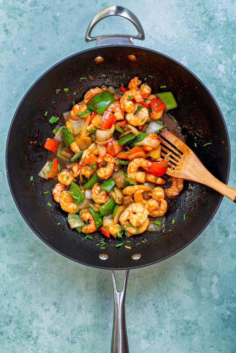 A wok filled with stir-fried shrimp, red and green bell peppers, onions, and herbs, with a wooden spatula resting inside. The background is a light blue textured surface.