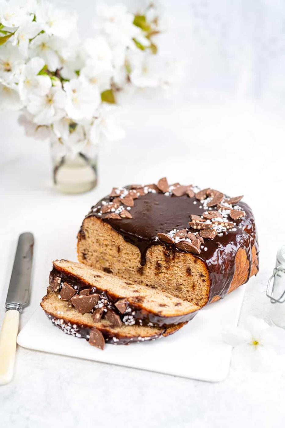 A chocolate-glazed loaf cake, reminiscent of slow cooker banana bread, with chocolate pieces and sugar sprinkles on top, is partially sliced on a white board next to a knife, with white flowers in a vase in the background.