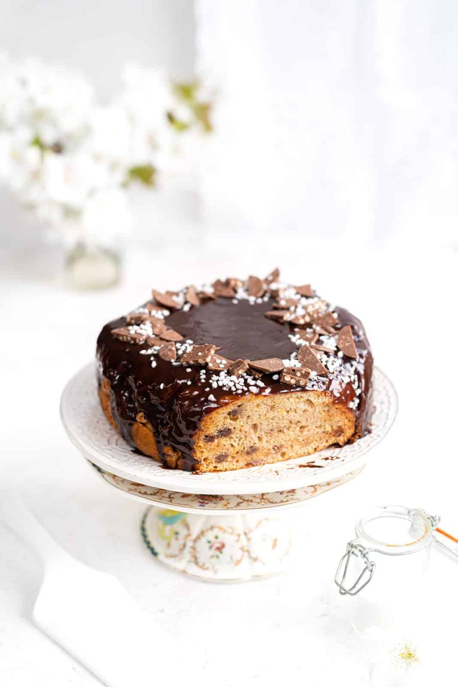 A round cake with chocolate glaze and chocolate pieces on top sits on a decorative cake stand, a slice missing—reminiscent of an indulgent 3-ingredient banana bread. White flowers and a glass jar blur in the background.