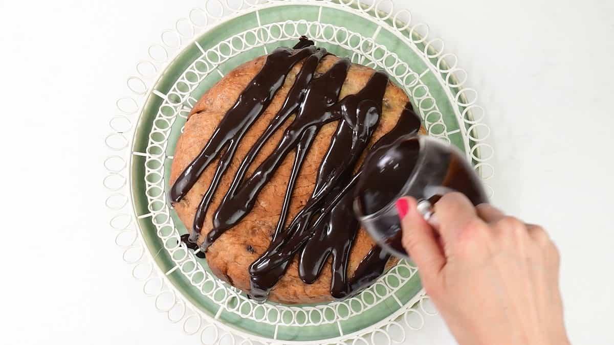 A hand pours chocolate sauce over a round cake, reminiscent of a slow cooker banana bread, placed on a green plate with a decorative white rim.
