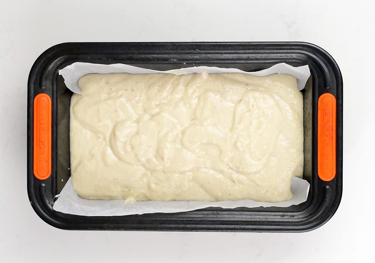 A black rectangular loaf pan lined with parchment paper is filled with creamy white batter for 3-ingredient banana bread, ready to be baked. The pan sits on a white surface.