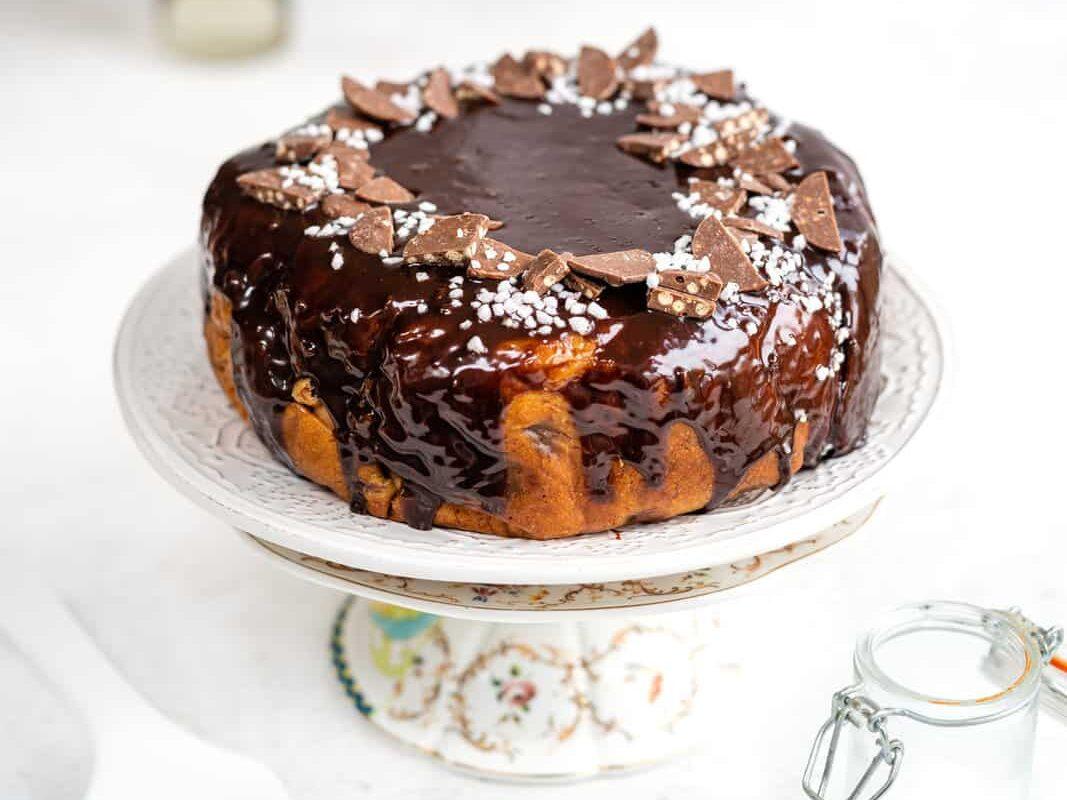 A round cake covered in glossy chocolate glaze, garnished with chocolate pieces and white sprinkles, sits on a decorative cake stand.