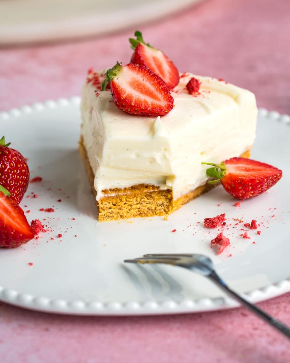 A slice of creamy three ingredient cheesecake topped with fresh strawberry halves sits on a white plate, with more strawberries and crumbs scattered around. A fork rests near the plate on a pink surface.