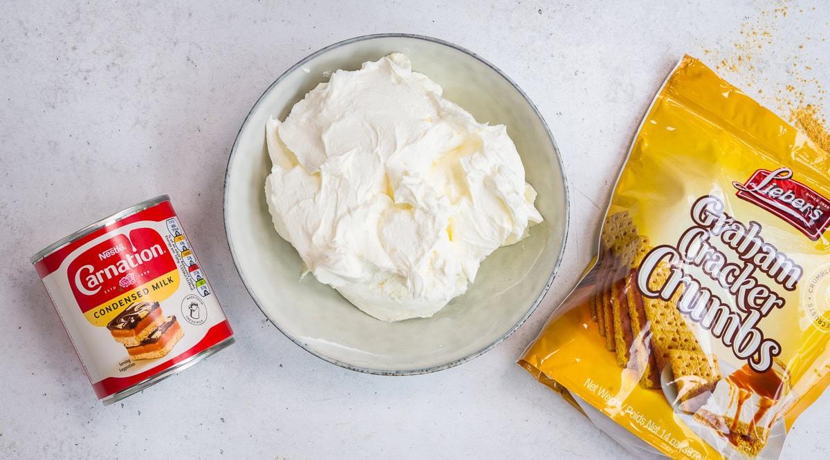 A can of Carnation condensed milk, a bowl of thick white cream cheese, and a bag of Lieber’s graham cracker crumbs on a white surface.