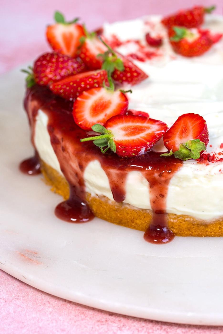 A no-bake cheesecake with a golden crust, topped with halved fresh strawberries and glossy strawberry sauce, sits on a white marble platter against a pink background.