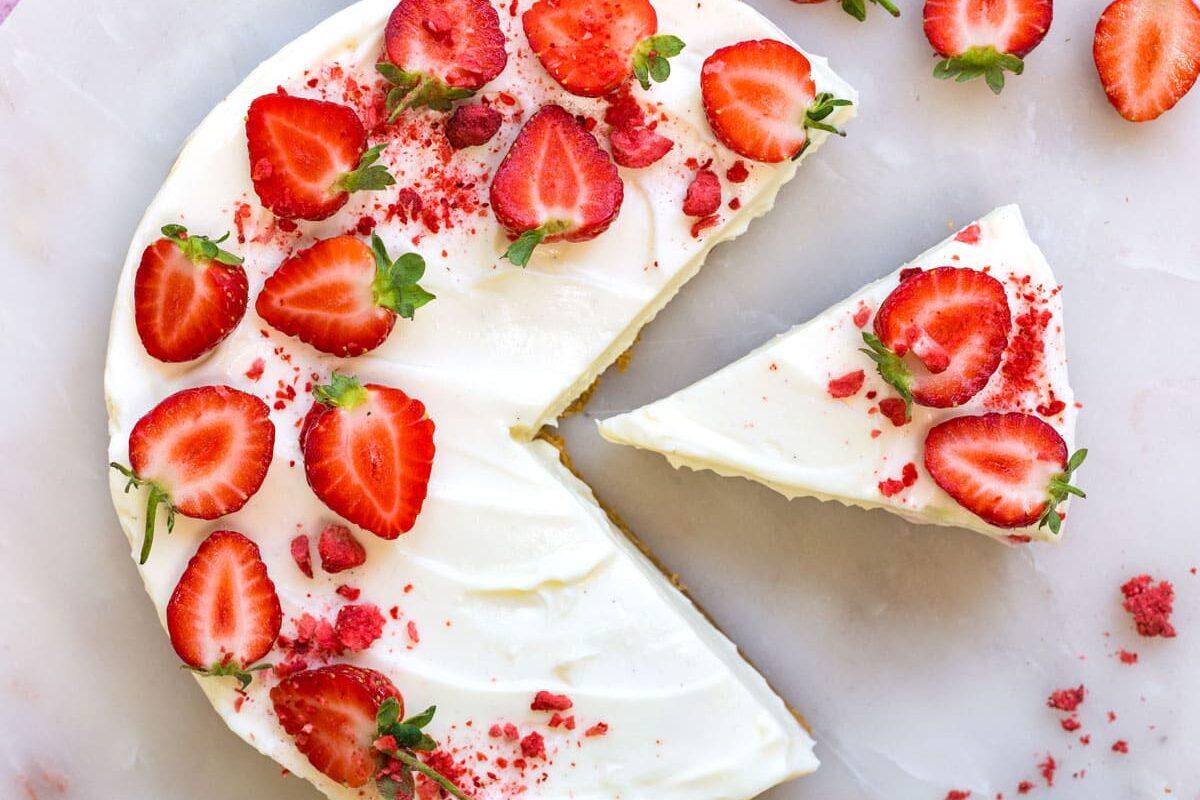 A round cake with white frosting topped with sliced strawberries, placed on a marble platter. One slice is cut and slightly pulled out. Whole and sliced strawberries are scattered nearby on a pink surface.