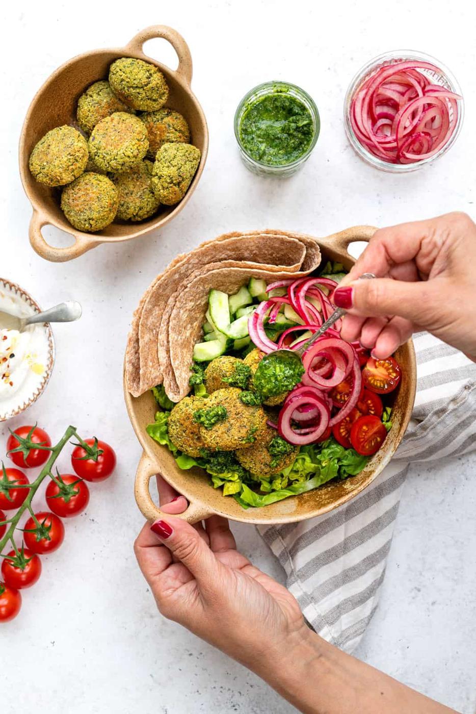 A bowl of falafel on greens with cucumber, tomato, pita, and pickled onions. A person adds pickled onions to this vibrant falafel recipe. Surrounding the bowl are more falafel, pickled onions, green sauce, yogurt, and cherry tomatoes on a vine.