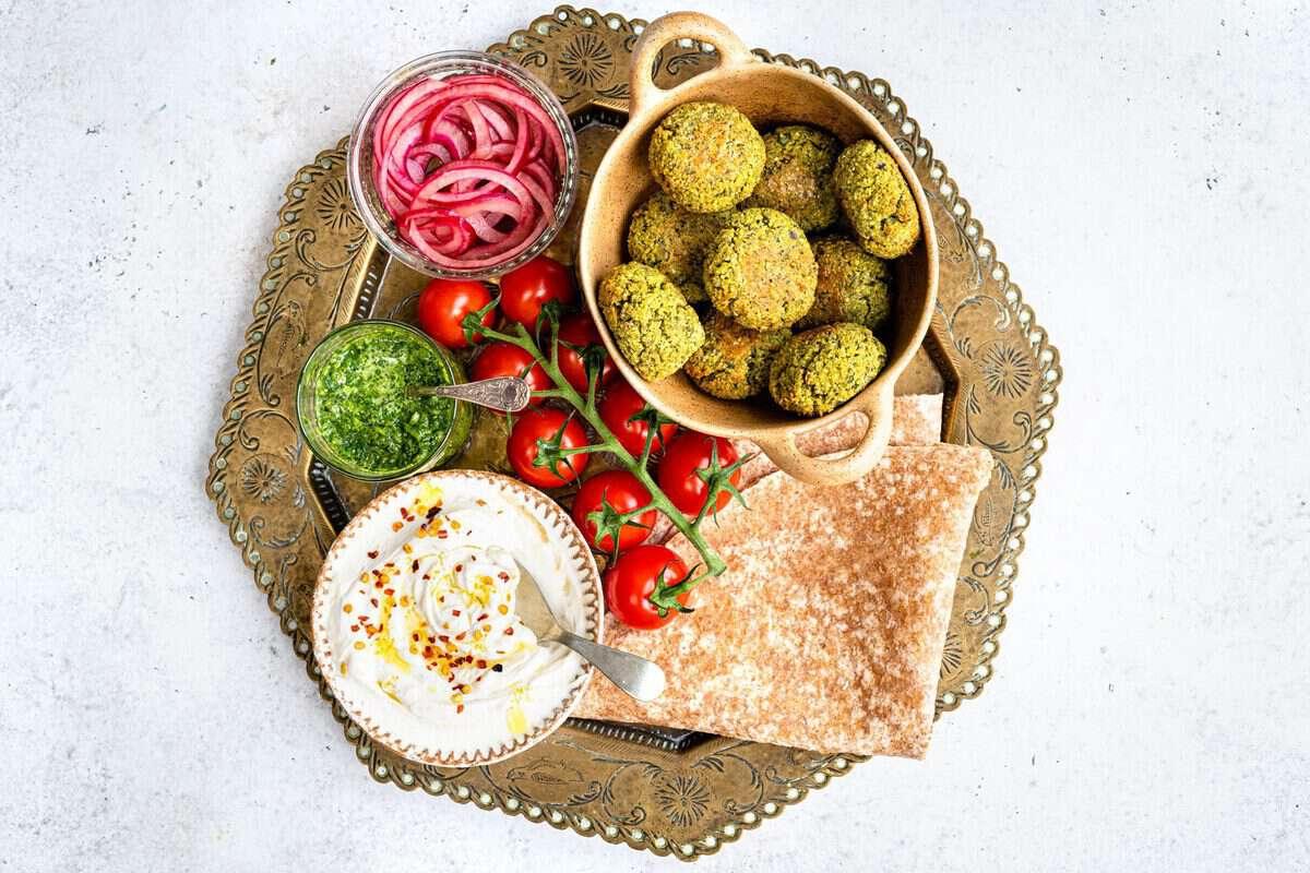 A decorative tray showcases a delicious falafel recipe with falafel in a bowl, pickled red onions in a jar, green sauce, vine tomatoes, flatbread, and yogurt with chili flakes on a light background.