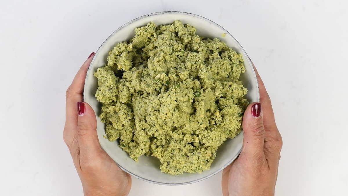 A pair of hands with red nail polish holds a bowl filled with a chunky, greenish mixture—perfect for your next falafel recipe—against a white background.