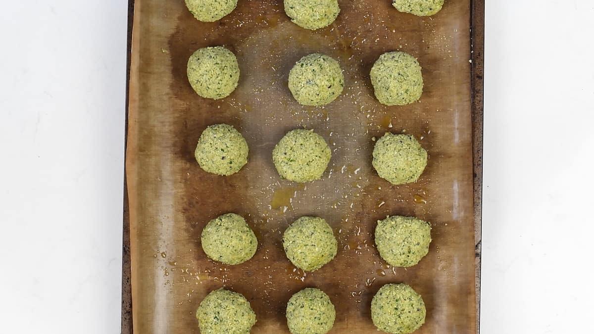A baking sheet lined with parchment paper holds fifteen evenly spaced, uncooked falafel balls made from a herbed mixture. This simple falafel recipe is finished with a sprinkle of olive oil and fresh herbs on top.