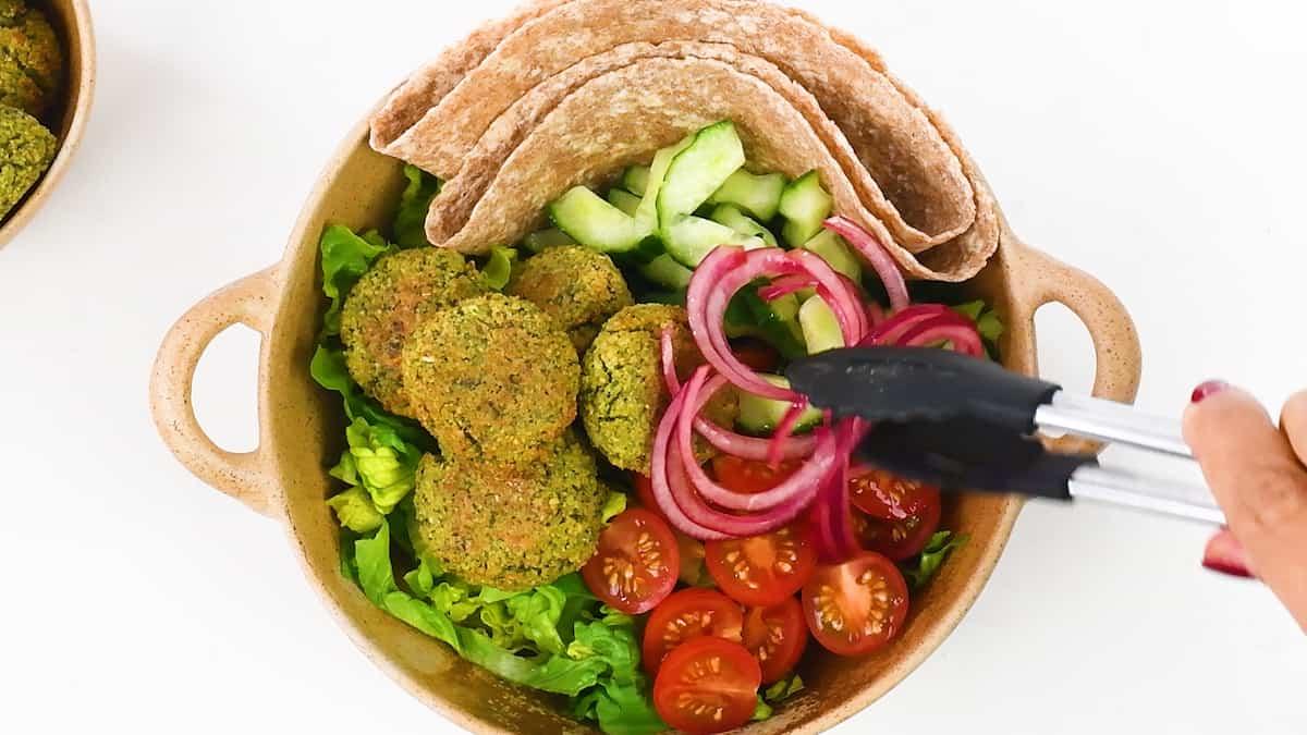 A bowl filled with a delicious falafel recipe, sliced cucumbers, cherry tomatoes, lettuce, pita bread, and pickled red onions being placed with tongs, arranged on a white background.