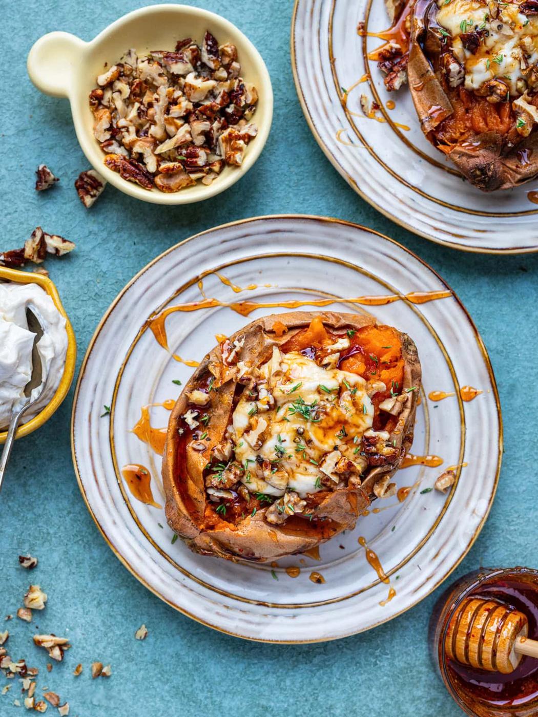 An air fryer baked sweet potato topped with whipped cheese, chopped pecans, and a drizzle of hot honey sits on a plate. Nearby are bowls of whipped cheese, chopped pecans, and honey. Another plated sweet potato is partially visible.