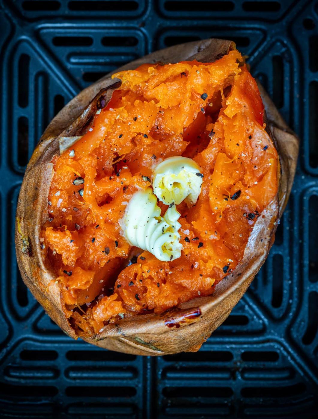 A baked sweet potato in topped with two curls of butter and a sprinkle of black pepper, sits in an air fryer basket.