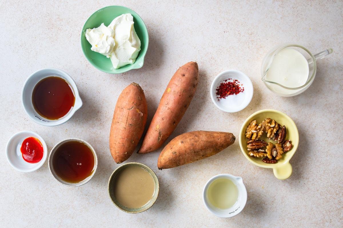 Three raw sweet potatoes surrounded by small bowls containing maple syrup, tahini, sour cream, milk, nuts, oil, salt with chili flakes, and ketchup, all arranged on a light countertop.