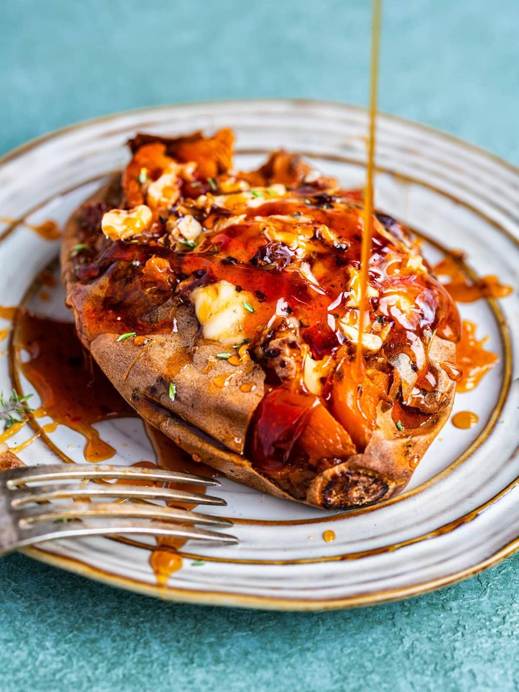 A baked sweet potato on a plate, topped with melting butter and herbs, with syrup being drizzled over it. A fork rests beside the potato on the plate.