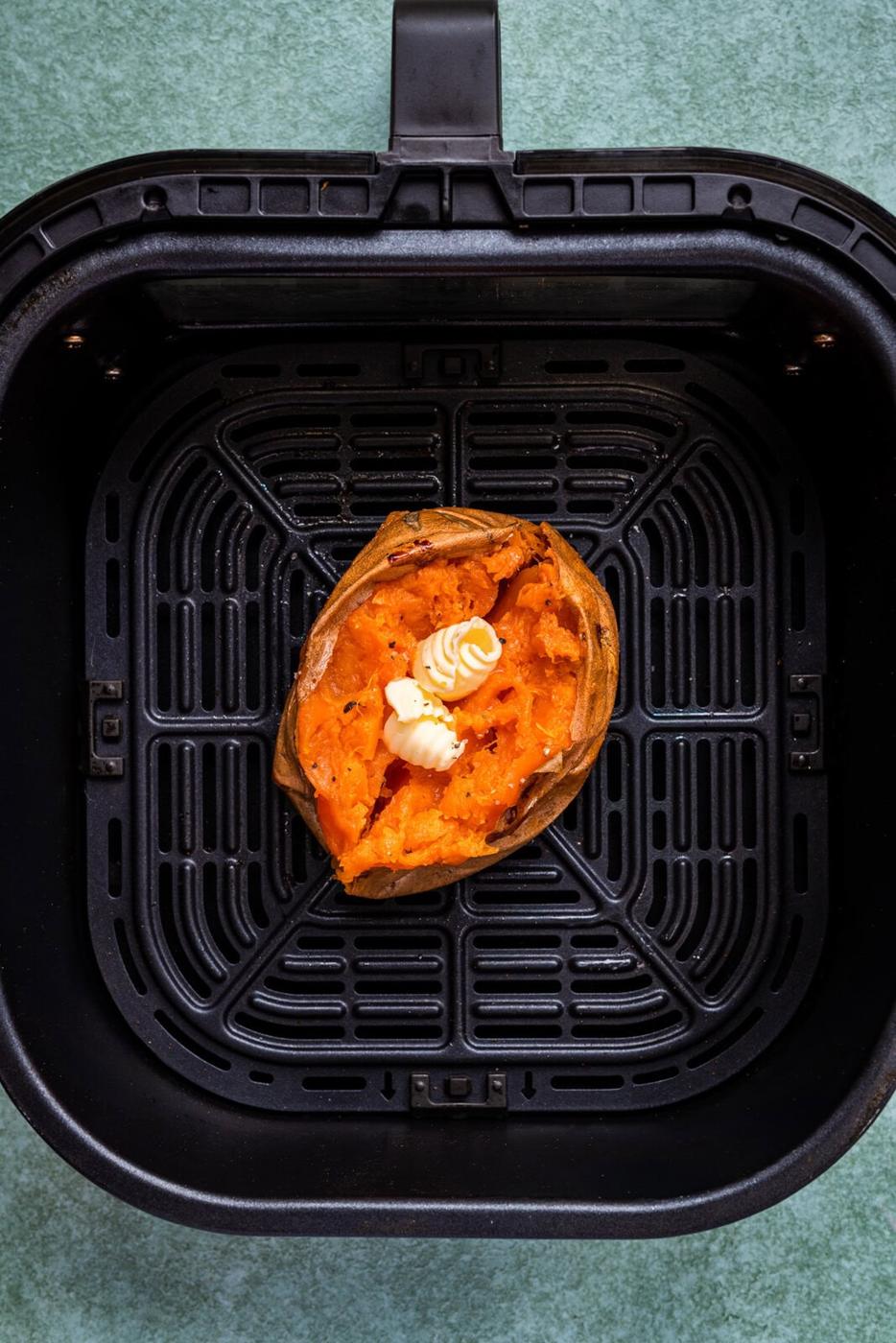 A baked sweet potato topped with a pat of butter sits in the basket of an air fryer against a green background.