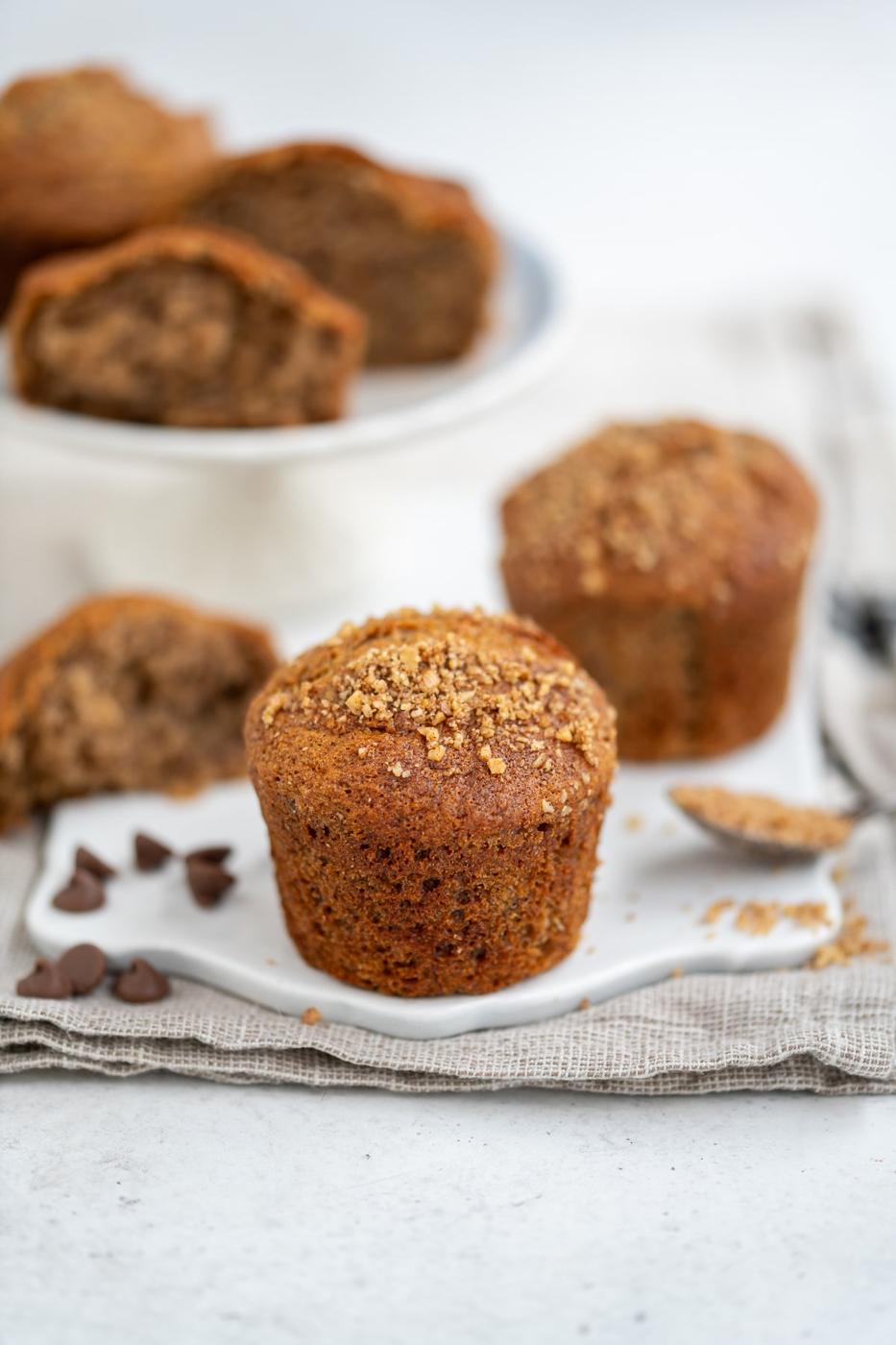 A close-up of Banana Bread Muffins topped with crumble on a white plate, with more muffins and chocolate chips in the background on a light textured surface—perfect for making in your Air Fryer.