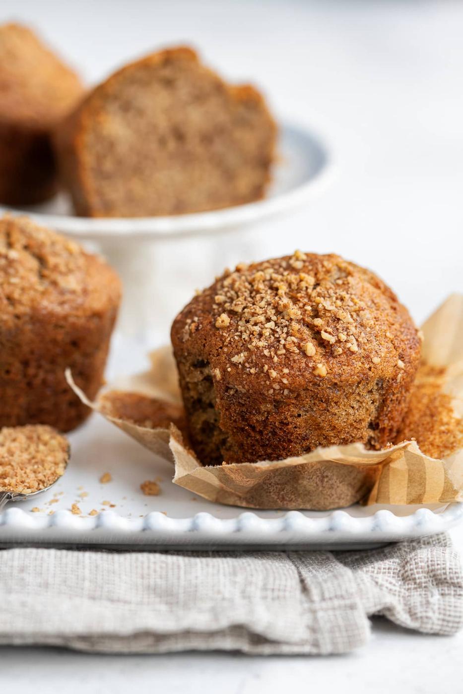 A close-up of a Banana Bread Muffin with crumb topping, partially wrapped in brown parchment paper, sits on a white plate. More muffins and a linen cloth appear in the background, perfect for your next Air Fryer baking session.