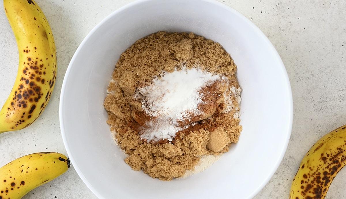 A white bowl filled with brown sugar, baking powder, and cinnamon sits on a light surface—perfect for making Banana Bread Muffins. Ripe bananas with brown spots are placed around the bowl, ready to be transformed in the Air Fryer.