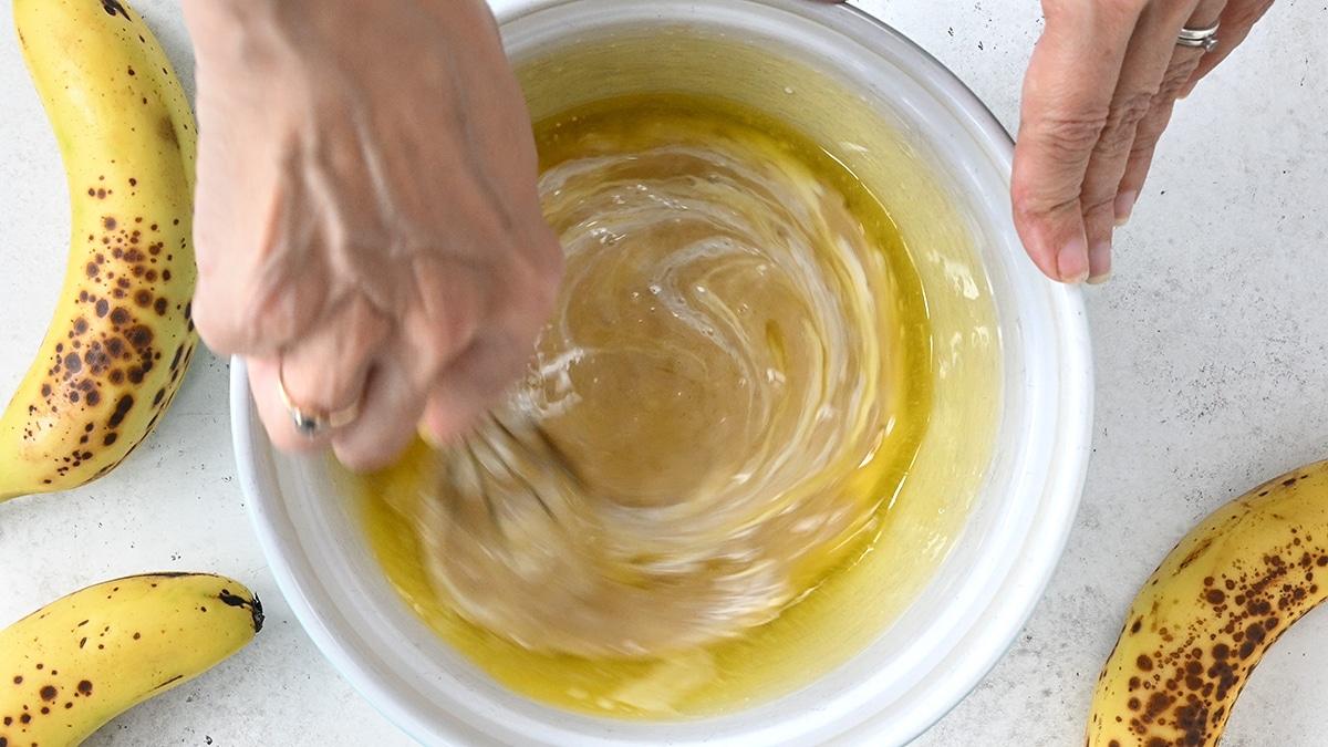 A person’s hands are mixing ingredients in a white bowl, surrounded by ripe bananas on a light countertop. The batter appears ready, perfect for making Banana Bread Muffins—perhaps even using an Air Fryer for a quick bake.