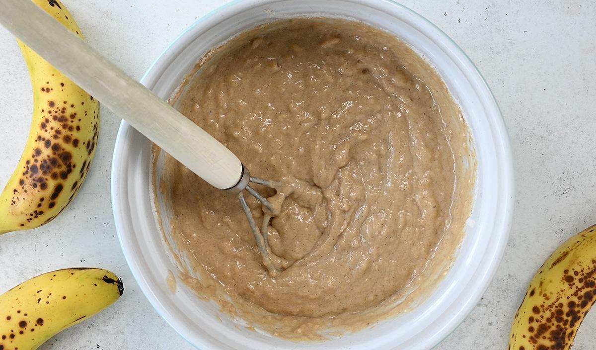 A mixing bowl filled with banana bread muffins batter sits on a light surface, surrounded by ripe bananas. A whisk rests in the bowl, ready to help you create delicious treats—perfect for baking in your air fryer.
