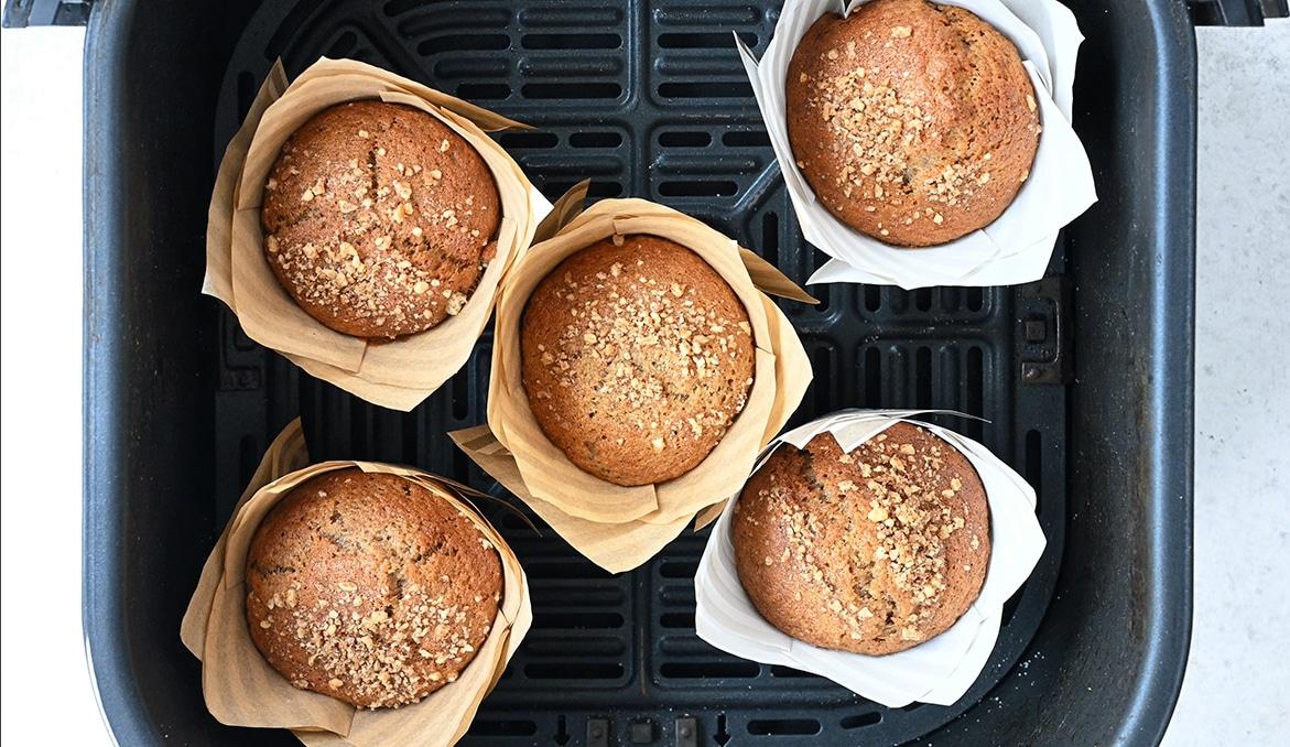 Five banana bread muffins in brown and white parchment paper cups are arranged in the basket of an air fryer. The muffins have a golden-brown top and are sprinkled with crushed nuts.