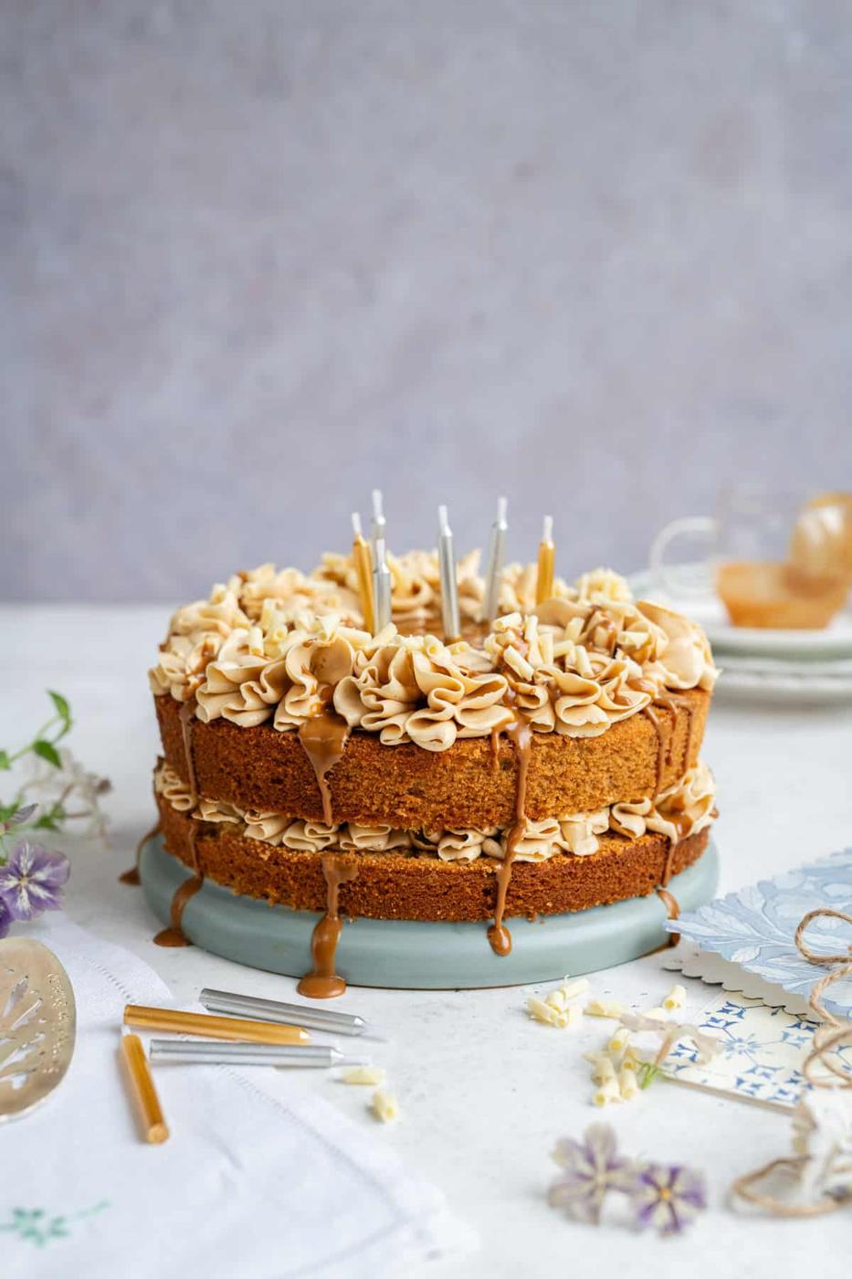 A two-layer cookie butter cake with decorative frosting, caramel drizzle, and silver birthday candles sits on a light blue cake stand. I