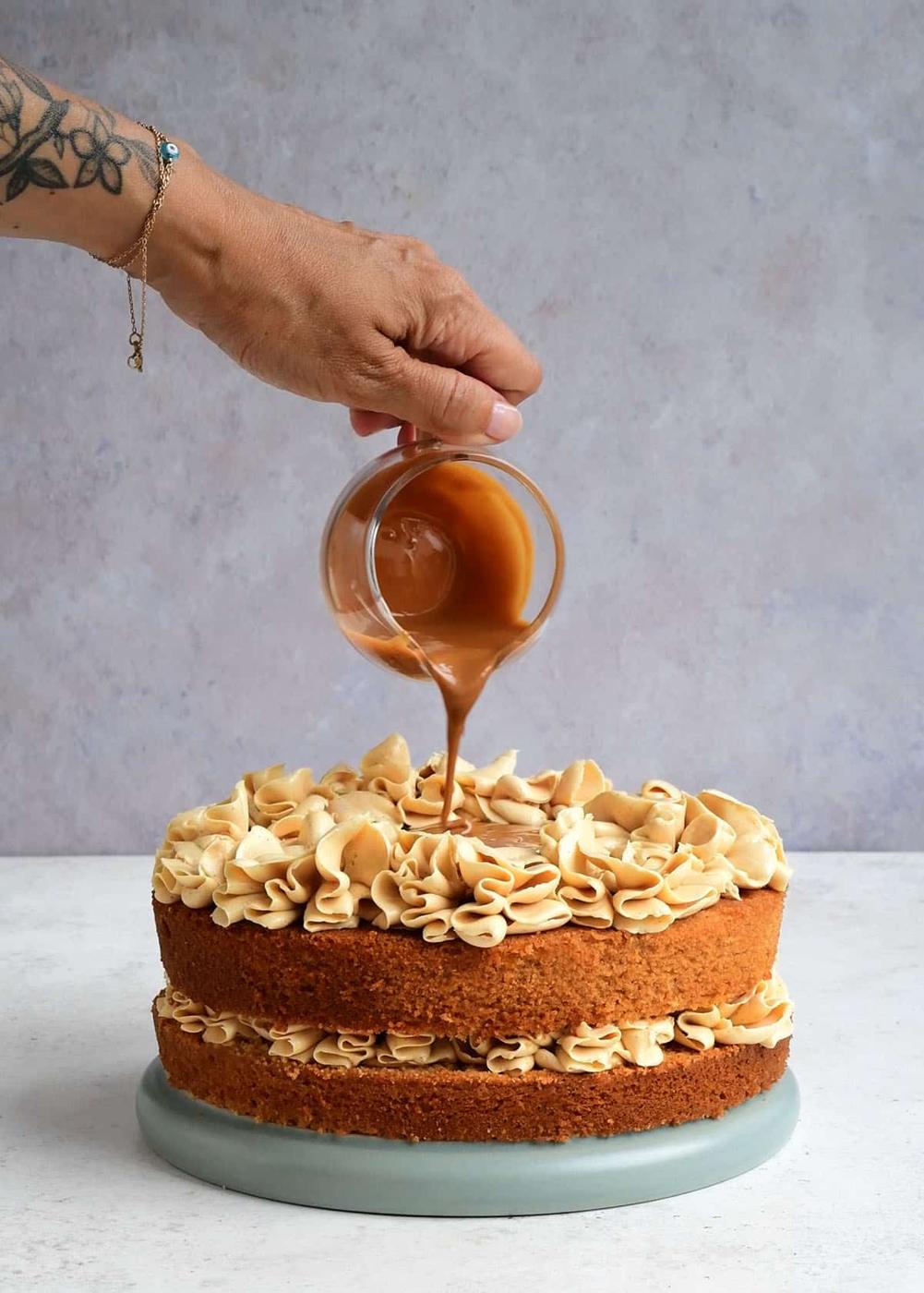 A hand with a tattoo pours caramel sauce from a glass cup onto an Air Fryer Biscoff Cake topped with swirls of frosting, sitting on a blue plate against a light background.