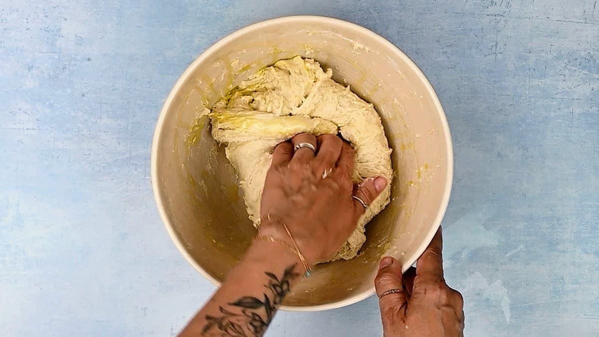 A tattooed hand kneads dough for air fryer bread in a large beige mixing bowl on a light blue textured surface.