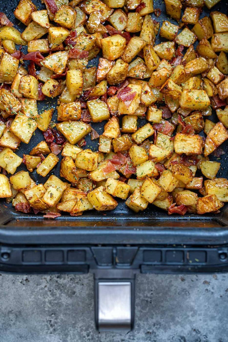 Golden roasted Air Fryer Breakfast Potatoes with herbs and crispy bacon pieces are spread evenly in an air fryer basket on a gray countertop.