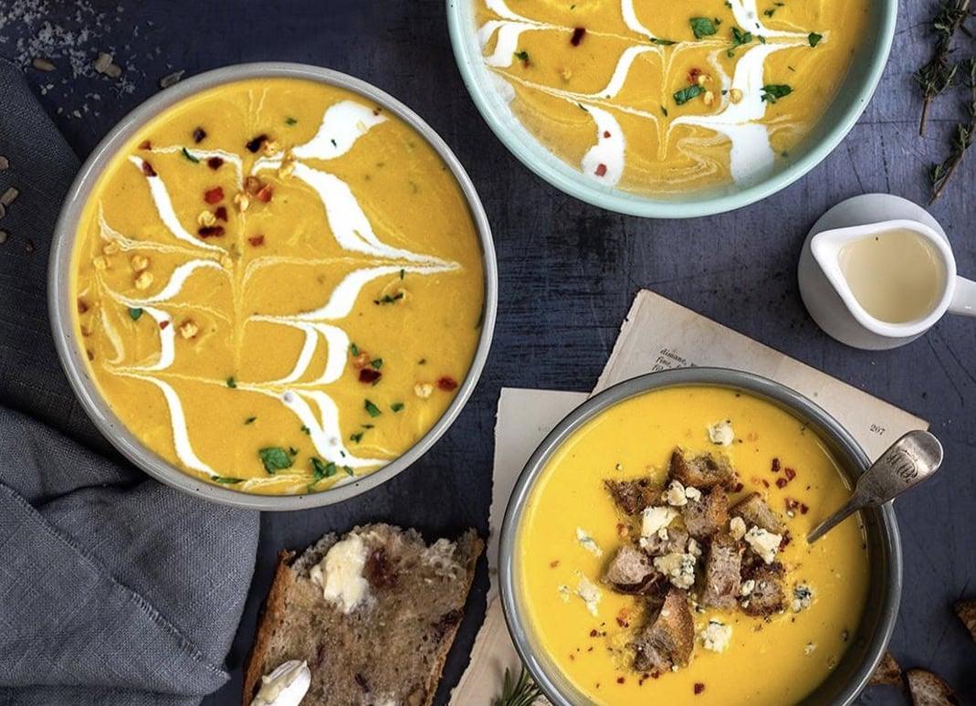 Three bowls of creamy orange Air Fryer Butternut Squash Soup, two with swirled white cream and herbs, and one topped with croutons and cheese. A slice of bread with butter and a small pitcher of cream sit nearby on a dark tabletop.