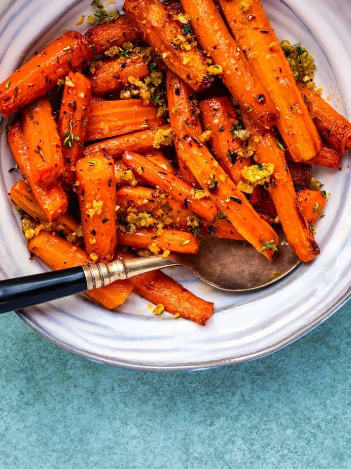 A plate of air fryer roasted carrots garnished with herbs and breadcrumbs, served with a spoon on a white dish against a light blue background.