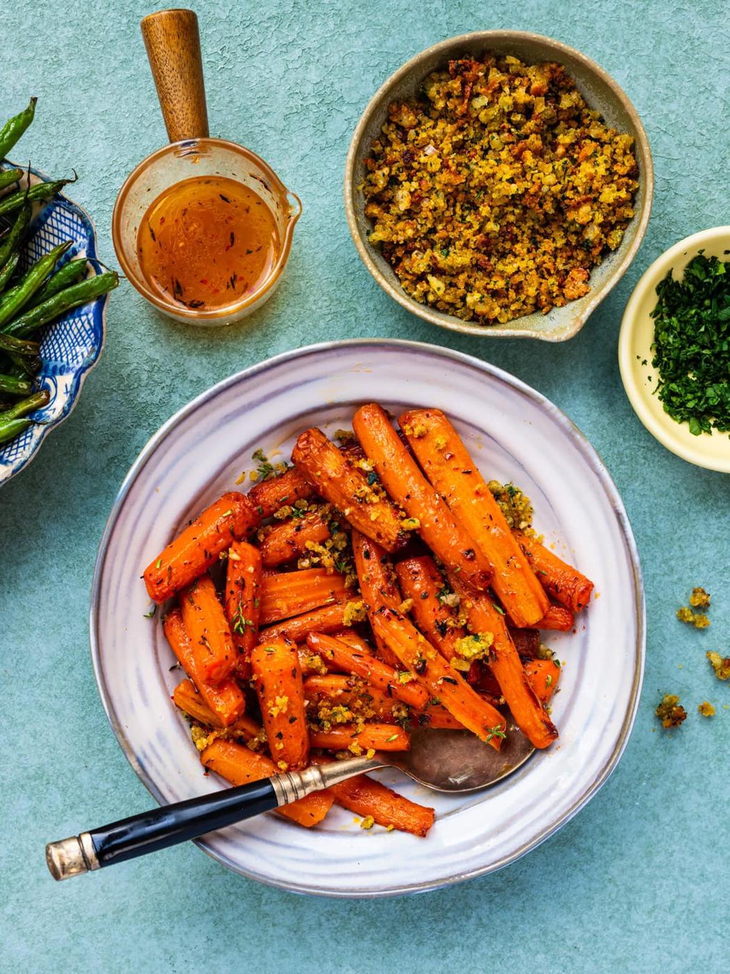 A bowl of air fryer roasted carrots garnished with herbs and crumbs sits on a table, surrounded by a dish of green beans, a bowl of crumbs, a bowl of chopped herbs, and a small pitcher of dressing. A spoon and fork rest in the carrot bowl.