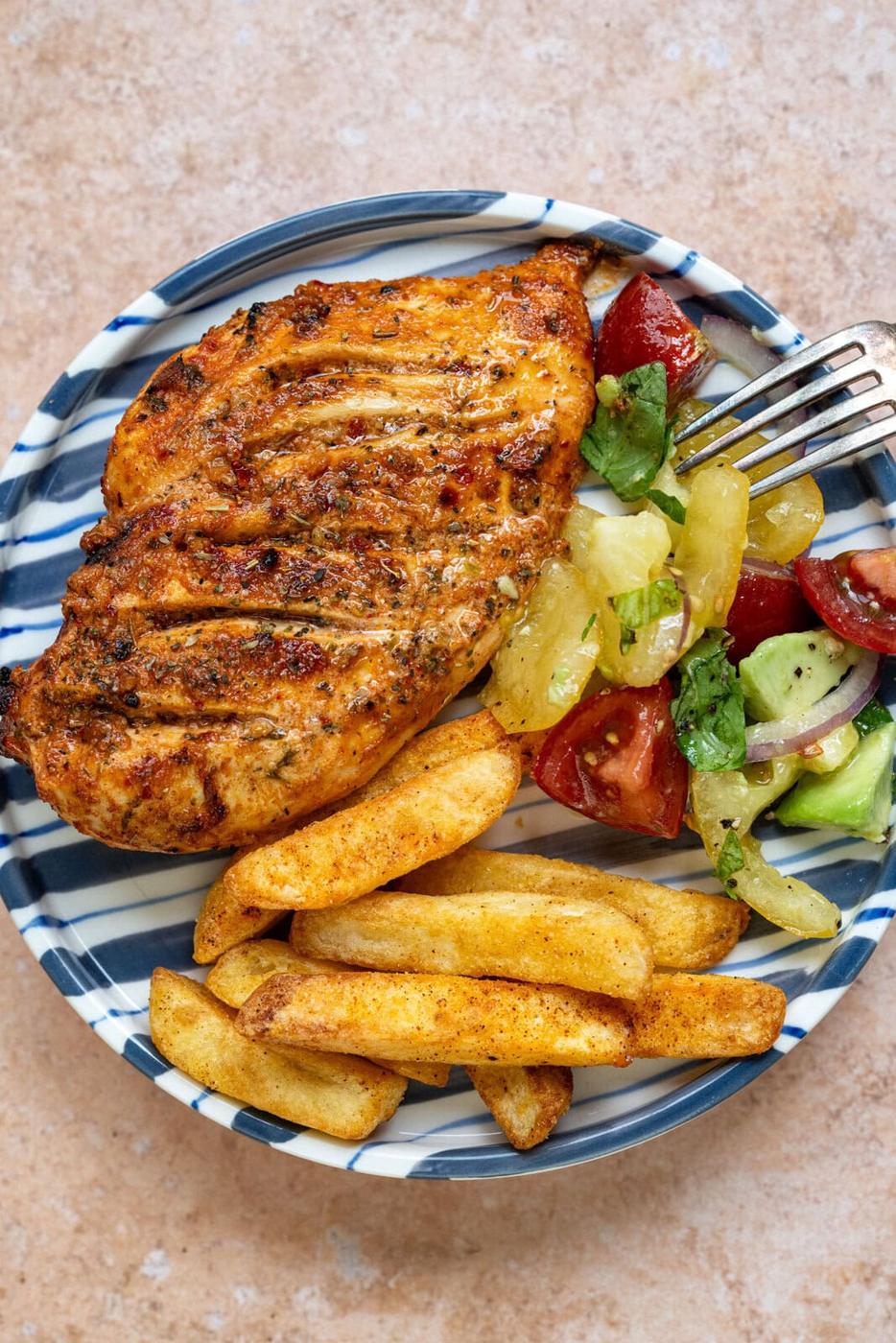 A plate with air fryer chicken breast, seasoned potato fries, and a fresh salad of tomatoes, cucumbers, and greens. A fork rests on the plate. The plate has a blue-and-white striped rim on a beige surface.