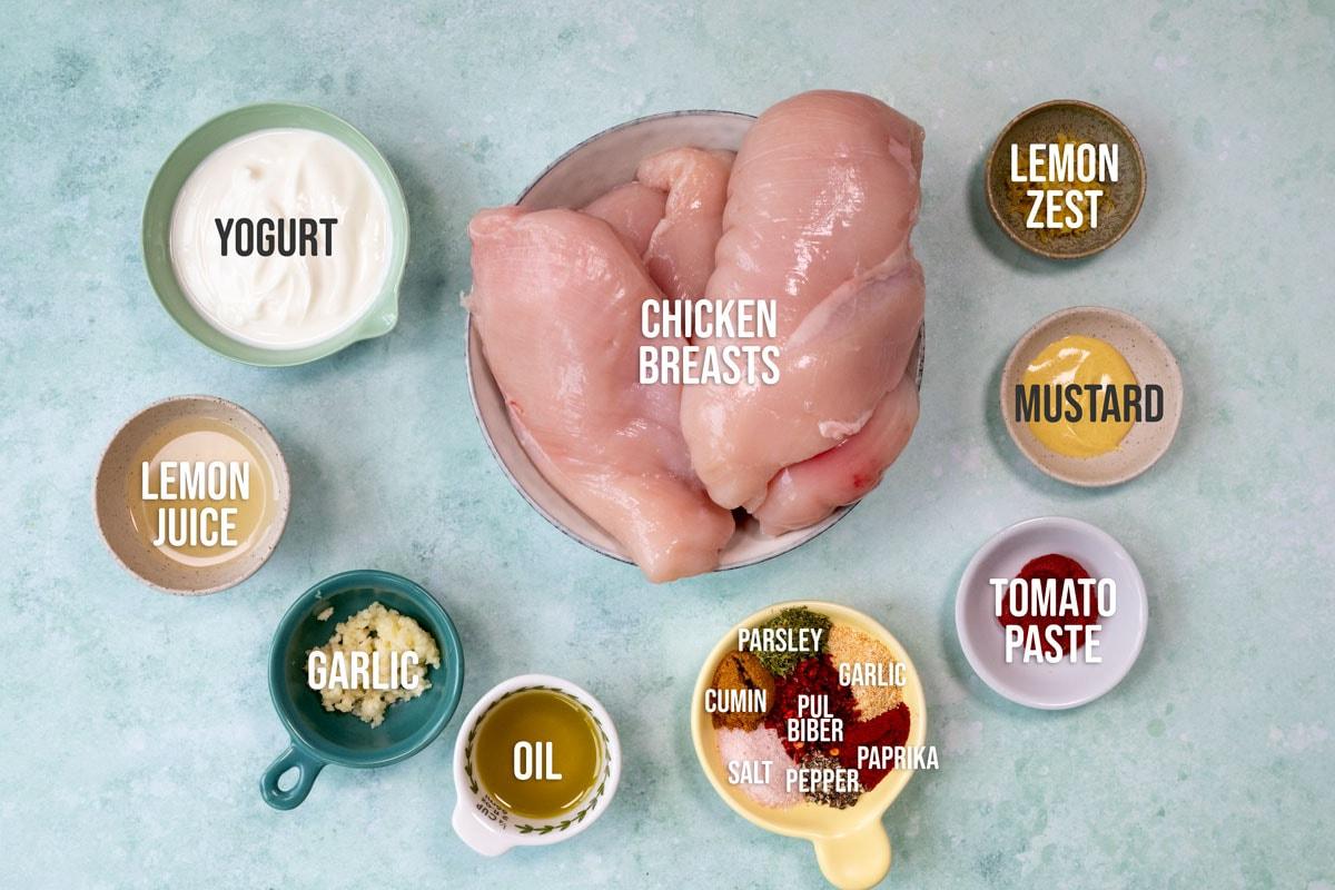Top-down view of raw chicken breasts in a bowl, surrounded by small bowls containing yogurt, lemon juice, garlic, oil, parsley, cumin, salt, pepper, Pul biber, paprika, lemon zest, mustard, and tomato paste.
