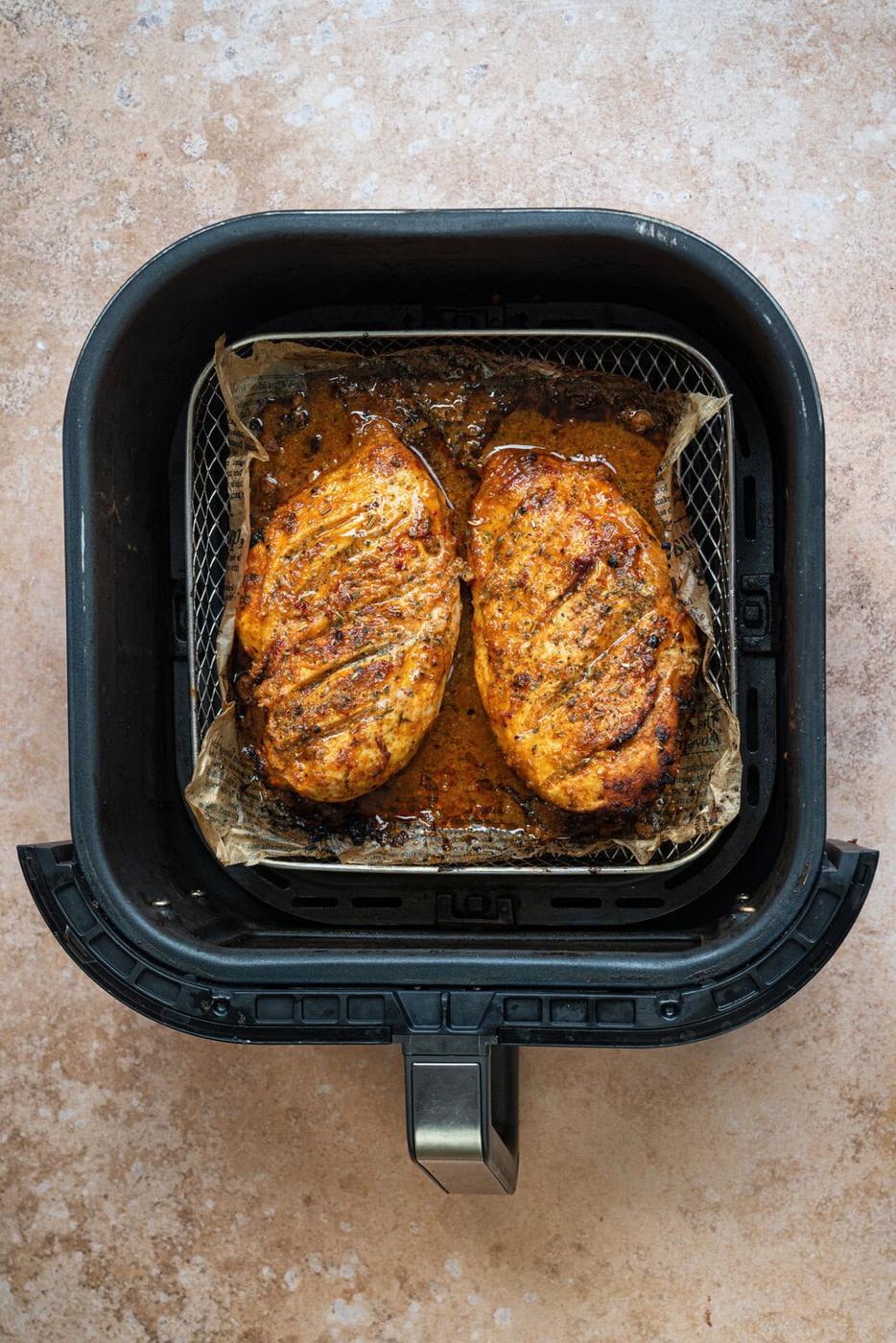Two seasoned, cooked chicken breasts in an air fryer basket lined with parchment paper, resting on a light brown countertop.