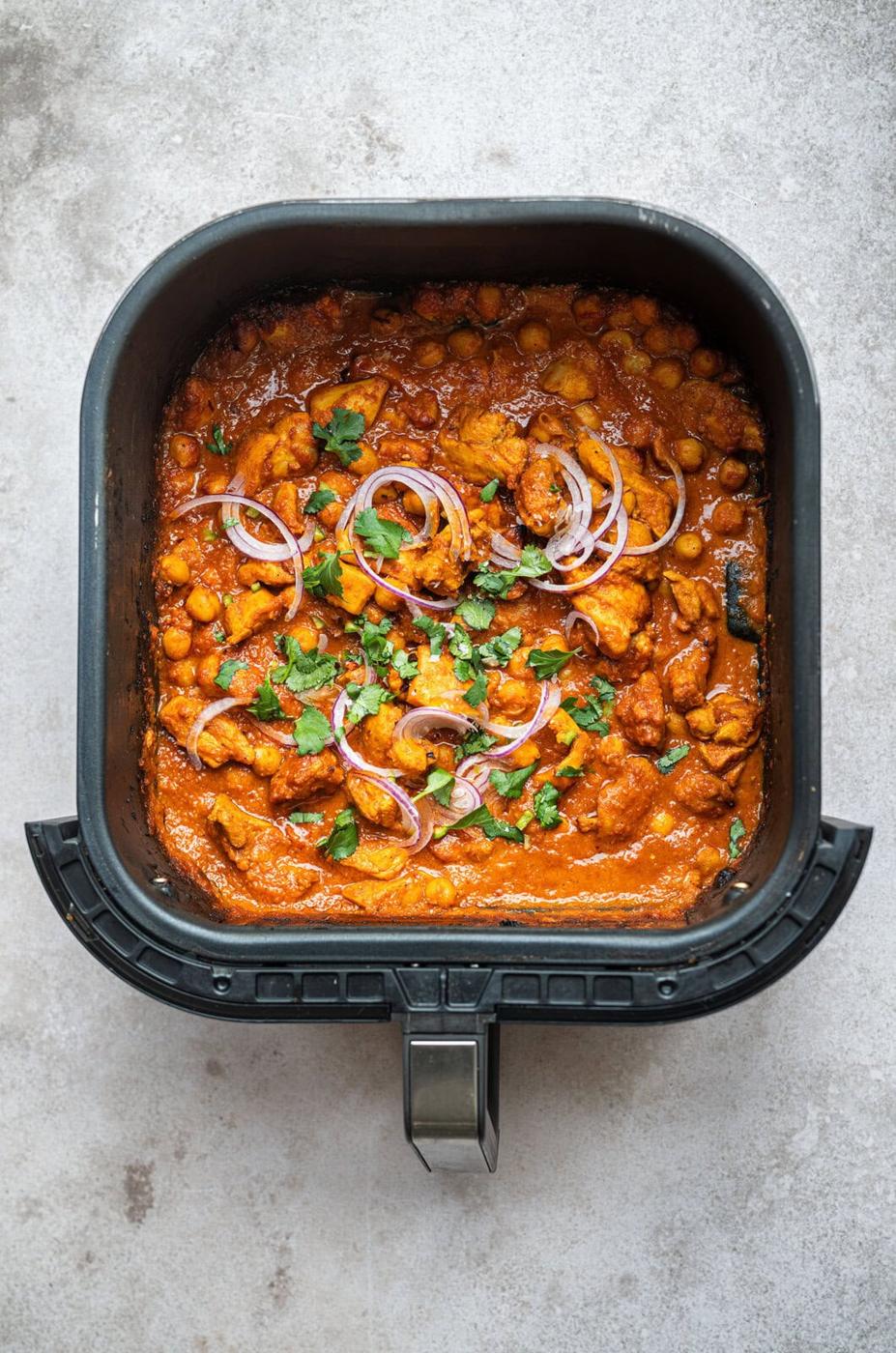 the basket of an air fryer filled with a vibrant curry containing pieces of chicken and chickpeas, garnished with thinly sliced red onions and fresh cilantro, placed on a light gray textured surface.