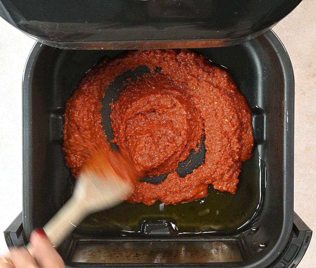 A hand stirring thick, red curry paste with a wooden spoon inside a black air fryer basket. The paste is spread in a circular pattern, contrasted against the black interior.