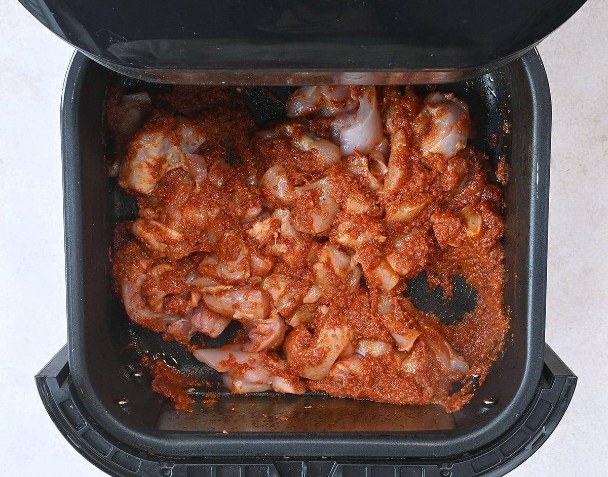 Pieces of raw chicken with a curry paste are placed inside an air fryer basket, ready for cooking. The marinade coats the chicken pieces evenly, and the basket is positioned on a light surface.