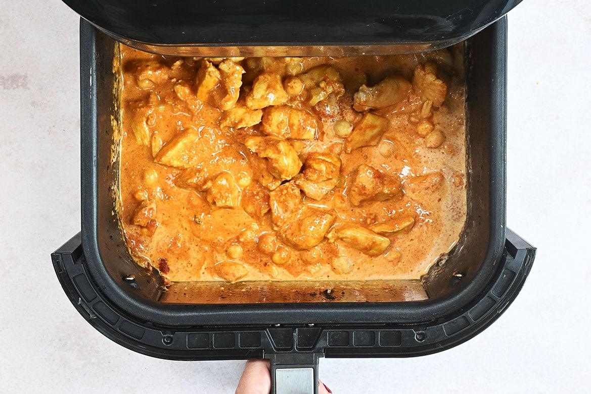 A hand pulling out an air fryer drawer containing a curry dish with chunks of chicken in a creamy, orange-colored curry sauce.