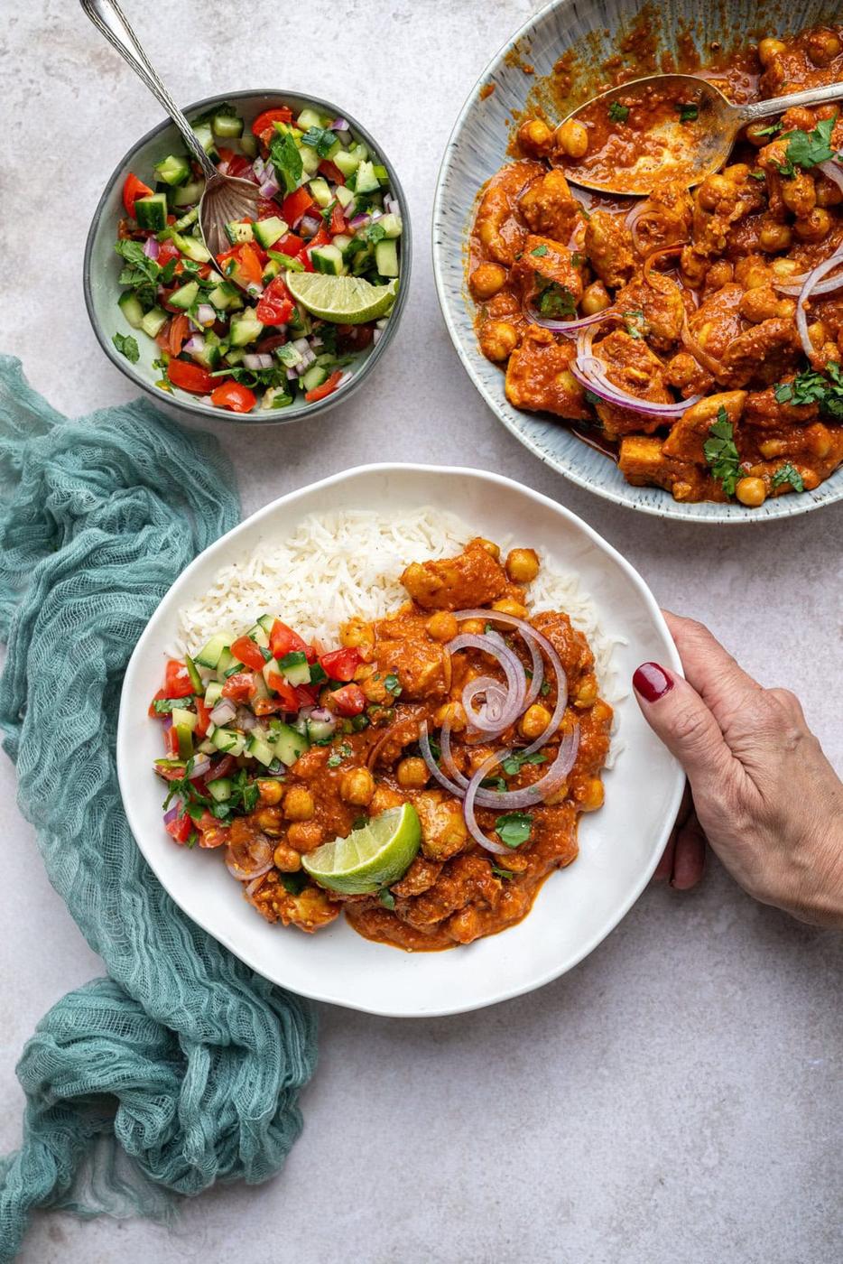 A hand holding a white plate of rice and chicken curry topped with onion slices and lime. A side of chopped salad is on the plate. Nearby, theres a ceramic bowl of curry and a small bowl of colorful salad on a grey surface with a green cloth.