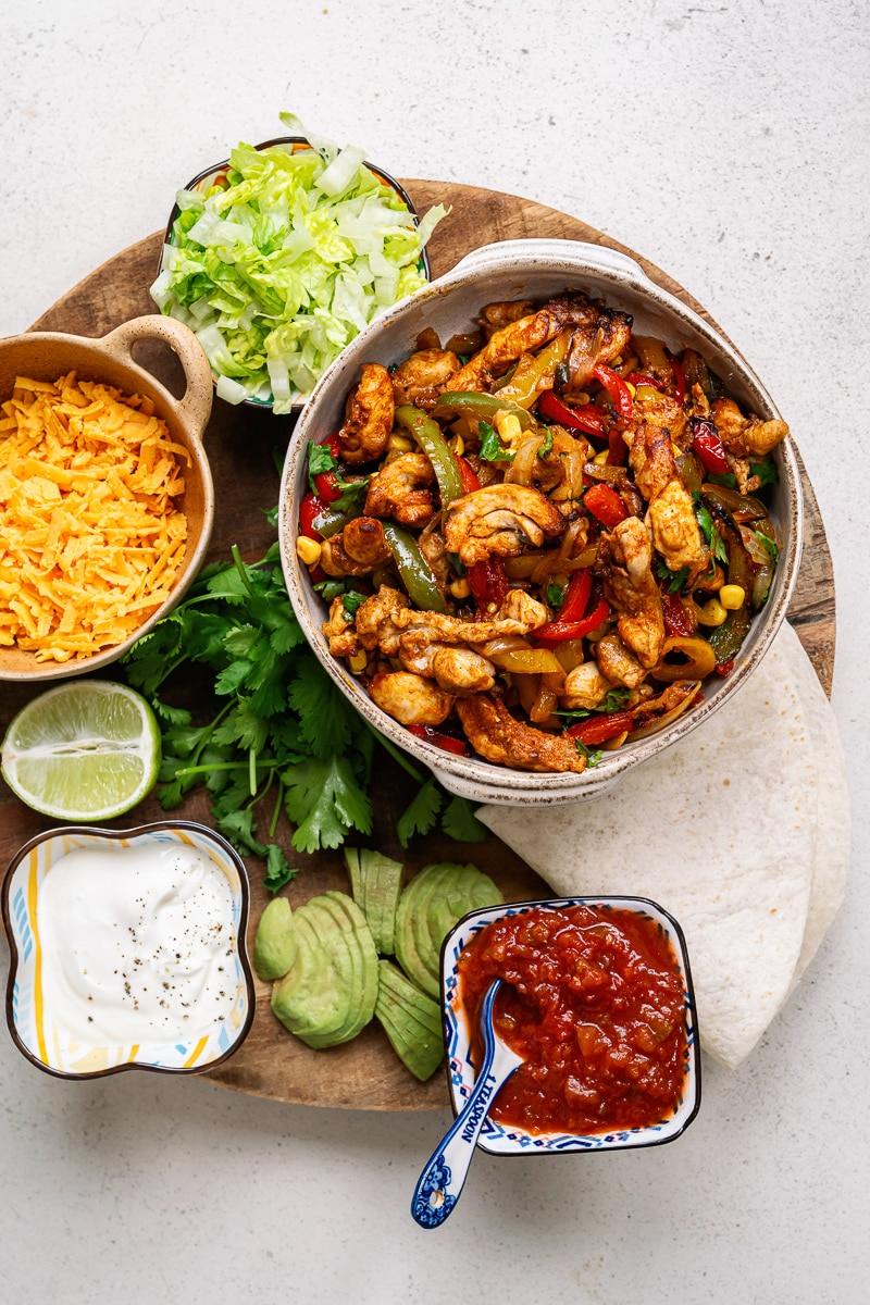 chicken fajitas in a bowl with grated cheese, avocado, salsa, coriander, lime, lettuce and sour cream arranged around it