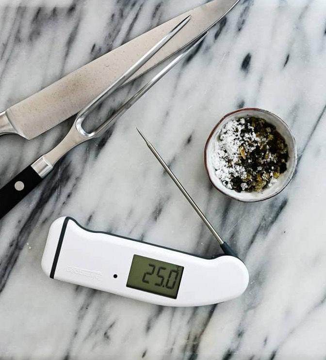 A digital thermometer displaying 25.0°C lies on a marble surface next to a large knife, a carving fork, and a small bowl of herbs and coarse salt—perfect for prepping Air Fryer Chicken Sharwama.