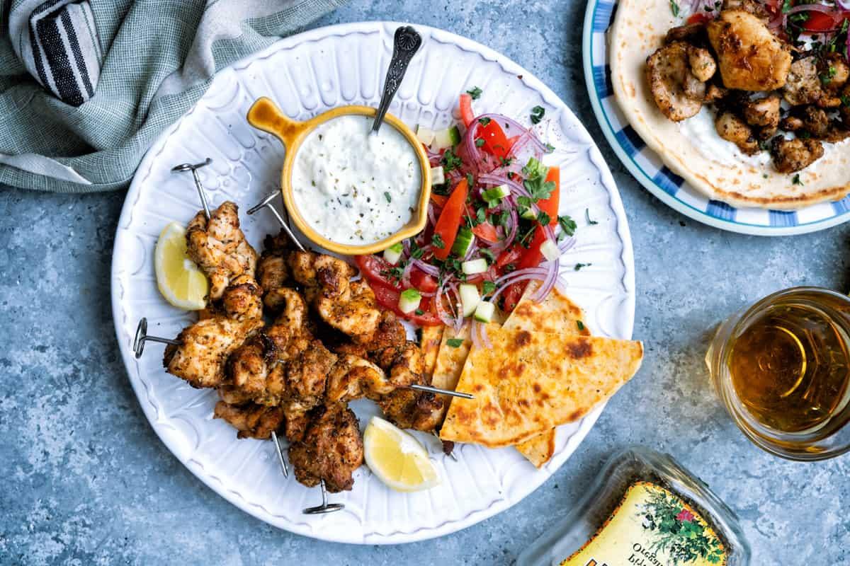 A plate of grilled chicken skewers with lemon wedges, a small bowl of creamy sauce, and a side of fresh salad with tomatoes and onions. Served alongside are pita slices.
