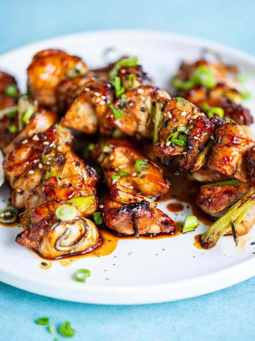 Air Fryer Japanese Yakitori chicken skewers glazed with a shiny sauce, garnished with sliced green onions, served on a white plate against a light blue background.
